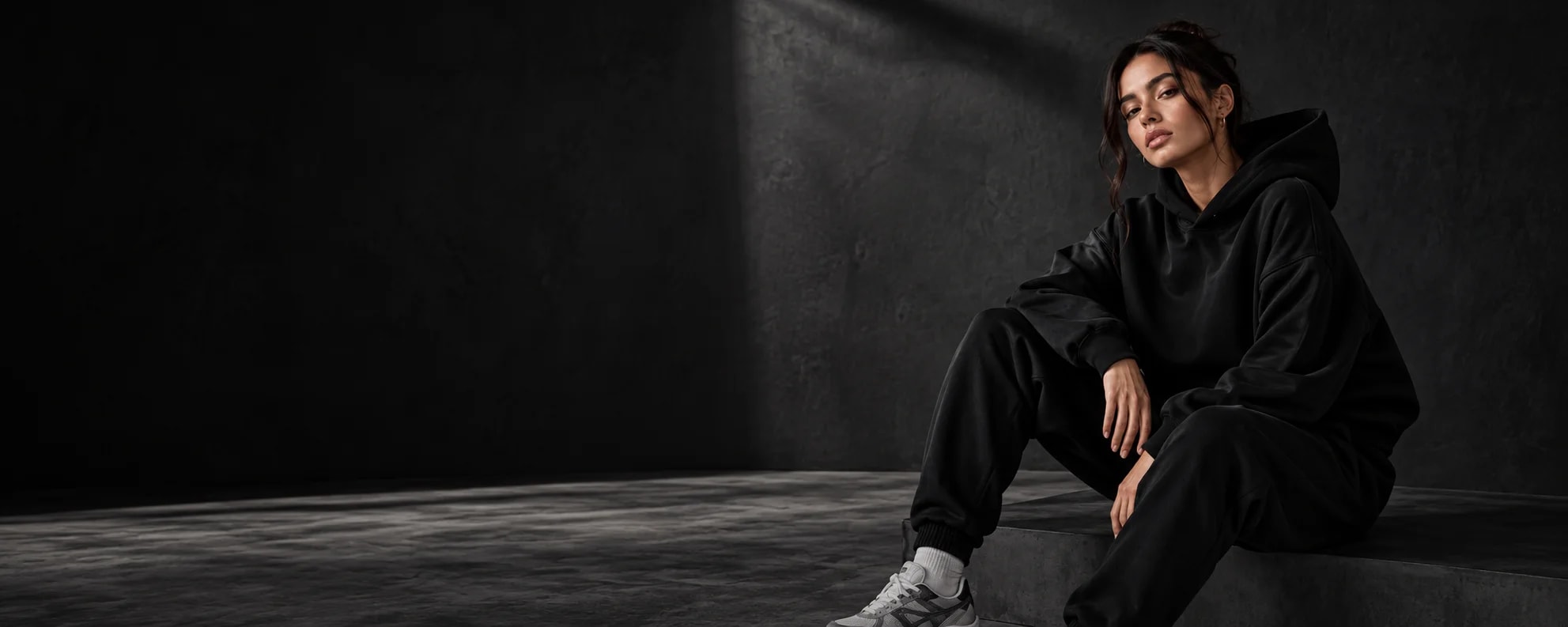 Woman seated in a black oversized hoodie and matching joggers with grey trainers